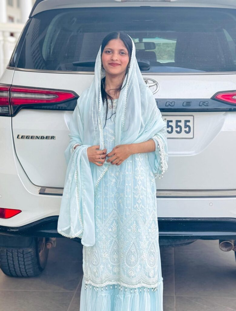 Woman in a light blue embroidered traditional outfit standing in front of a white SUV.